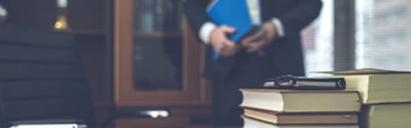 机の上に積み重なった書籍と、その後ろでスーツを着た男性が書類を持って立っている様子